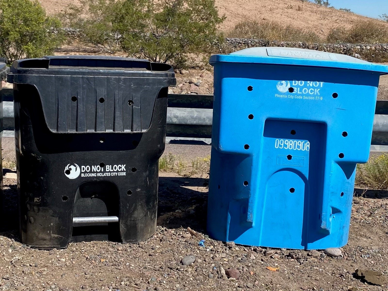 Two composters, one blue and one black, made from old solid waste containers. Each container has the bottom half sawed off and holes drilled throughout the sides to allow for aeration.