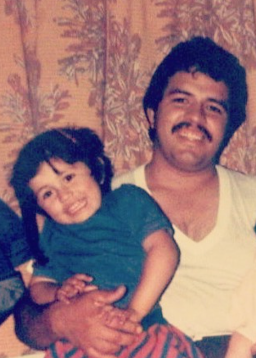 A faded vintage photo of a young Anna Hernandez as a child, smiling and being held by her father, who has a mustache and is wearing a white v-neck shirt, in front of a floral curtain backdrop.