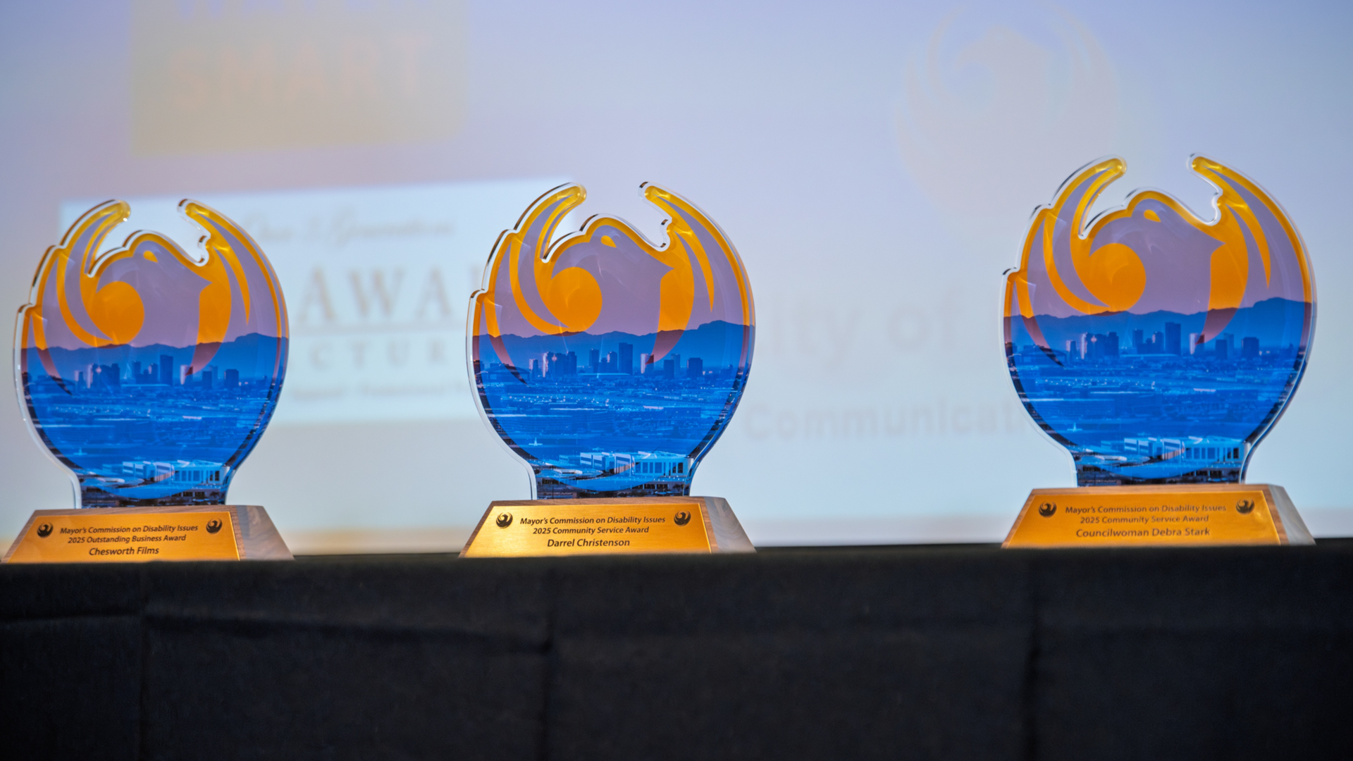 Image of three award statues in the shape of a Phoenix bird with a gold base. They are sitting on a black table with a glowing screen in the background. 