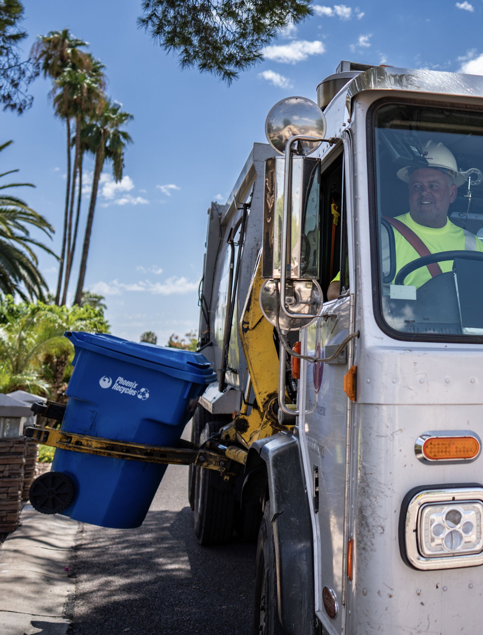 A solid waste equipment operator in a garbage truck uses a hydraulic lift to collect recyclables from a blue curbside bin.