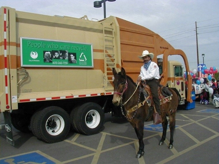 A man dressed as a cowboy sits atop a horse next to a tan Phoenix garbage truck.