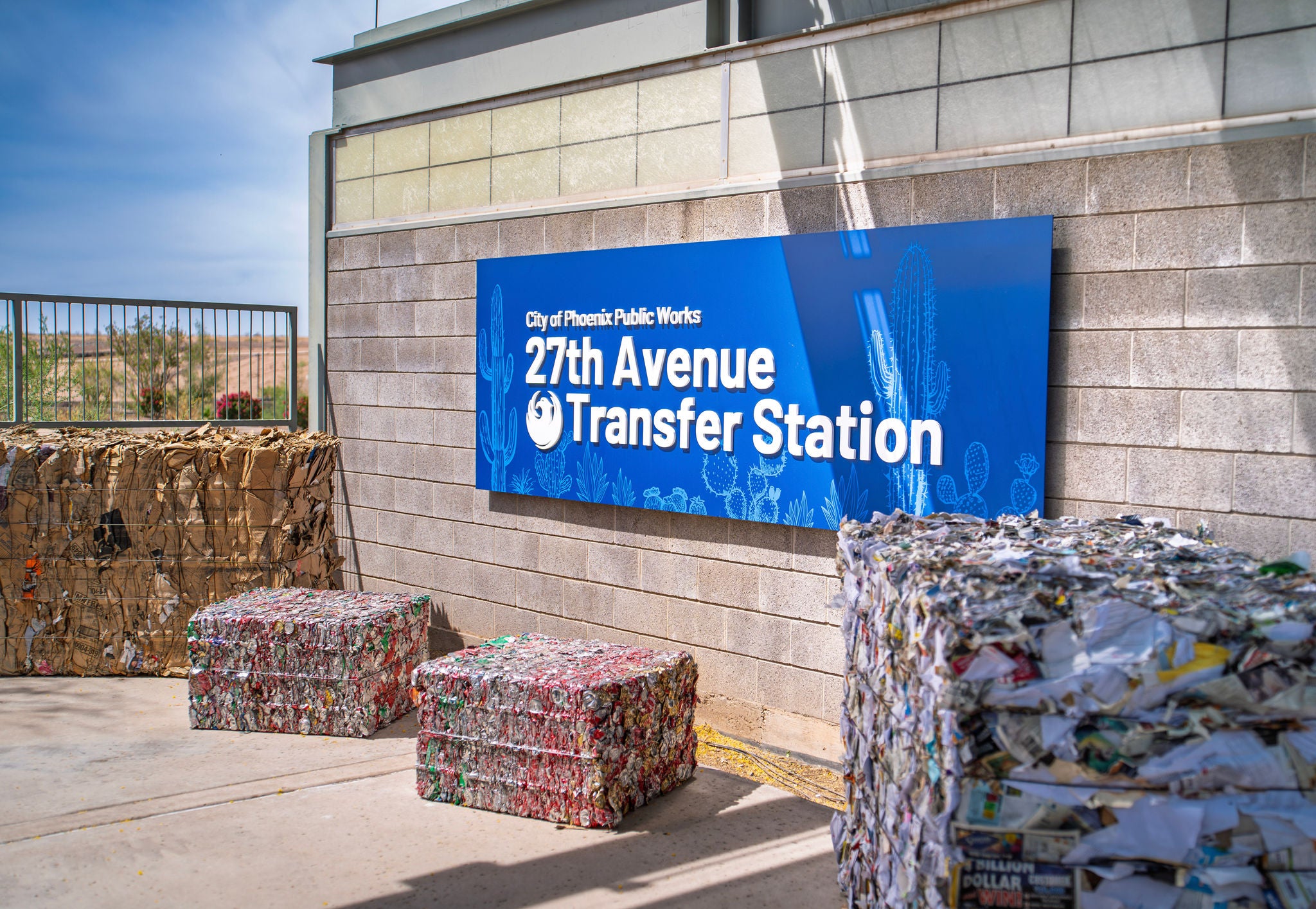 A blue sign on the side of the building that says 'City of Phoenix Public Works 27th Avenue Transfer Station.' There are bales of aluminum, cardboard, and paper sitting in front of the sign.