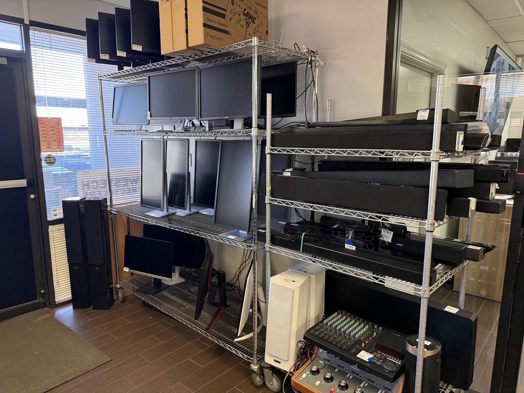 A room with two metal shelving units filled with electronics to be recycled -- monitors, computers, speakers, and switchboards. An external door is visible next to a window through which the sun is shining.