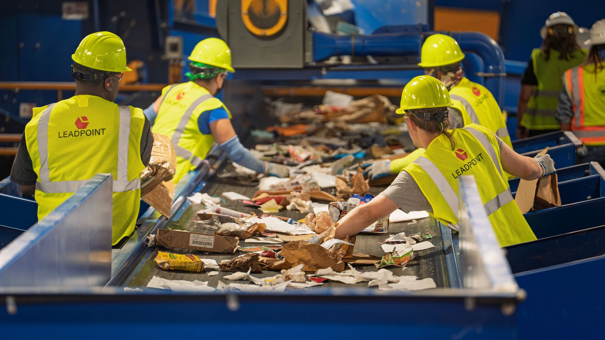 Workers in yellow hard hats and vests pull contaminants off of a conveyor belt of recyclables.