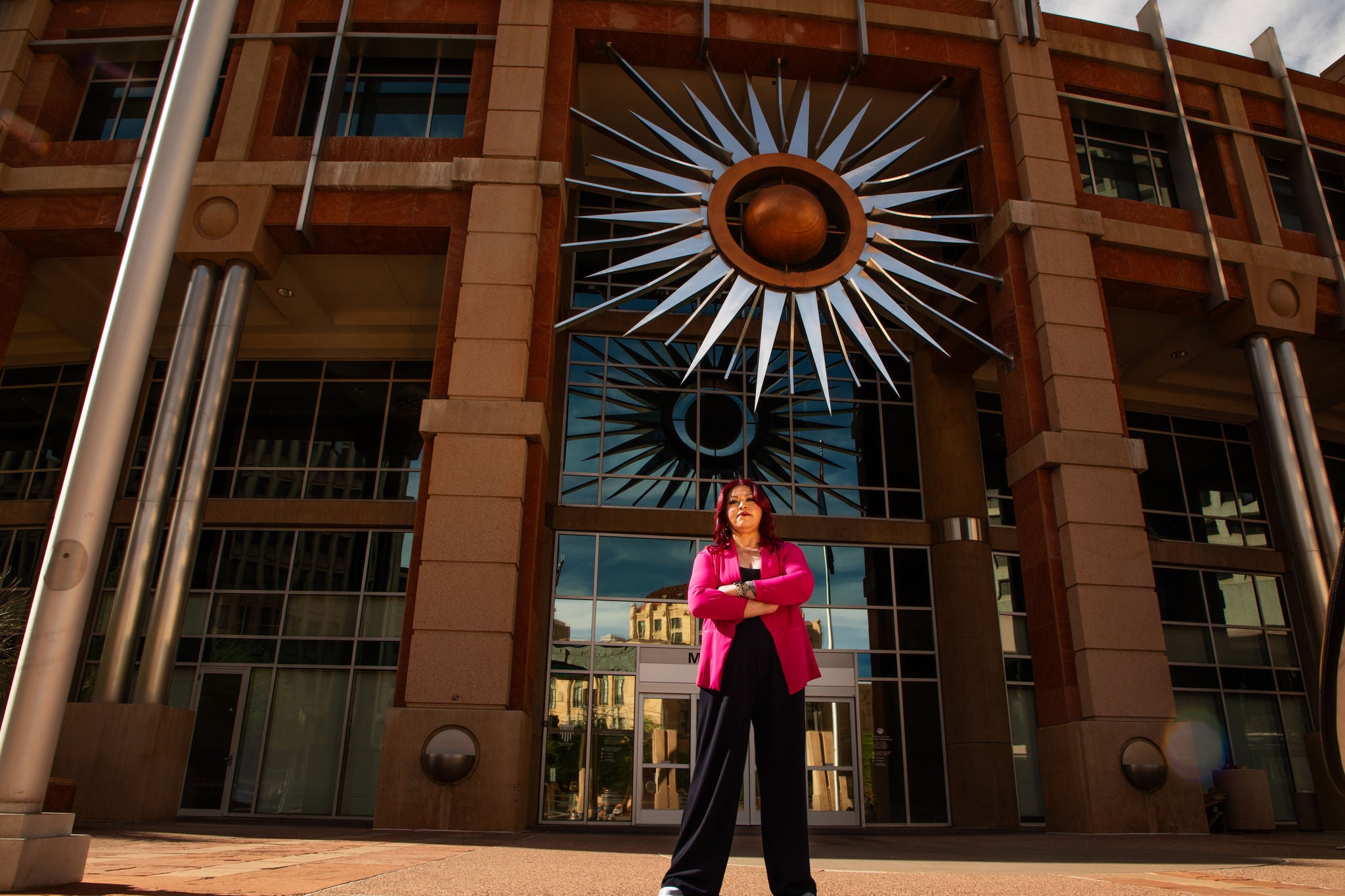 Councilmember Anna Hernandez stands with arms crossed in front of Phoenix City Hall, beneath the building’s striking sun sculpture. Dressed in a bold pink blazer, she exudes strength and determination, framed by the towering architecture and reflections of downtown Phoenix.