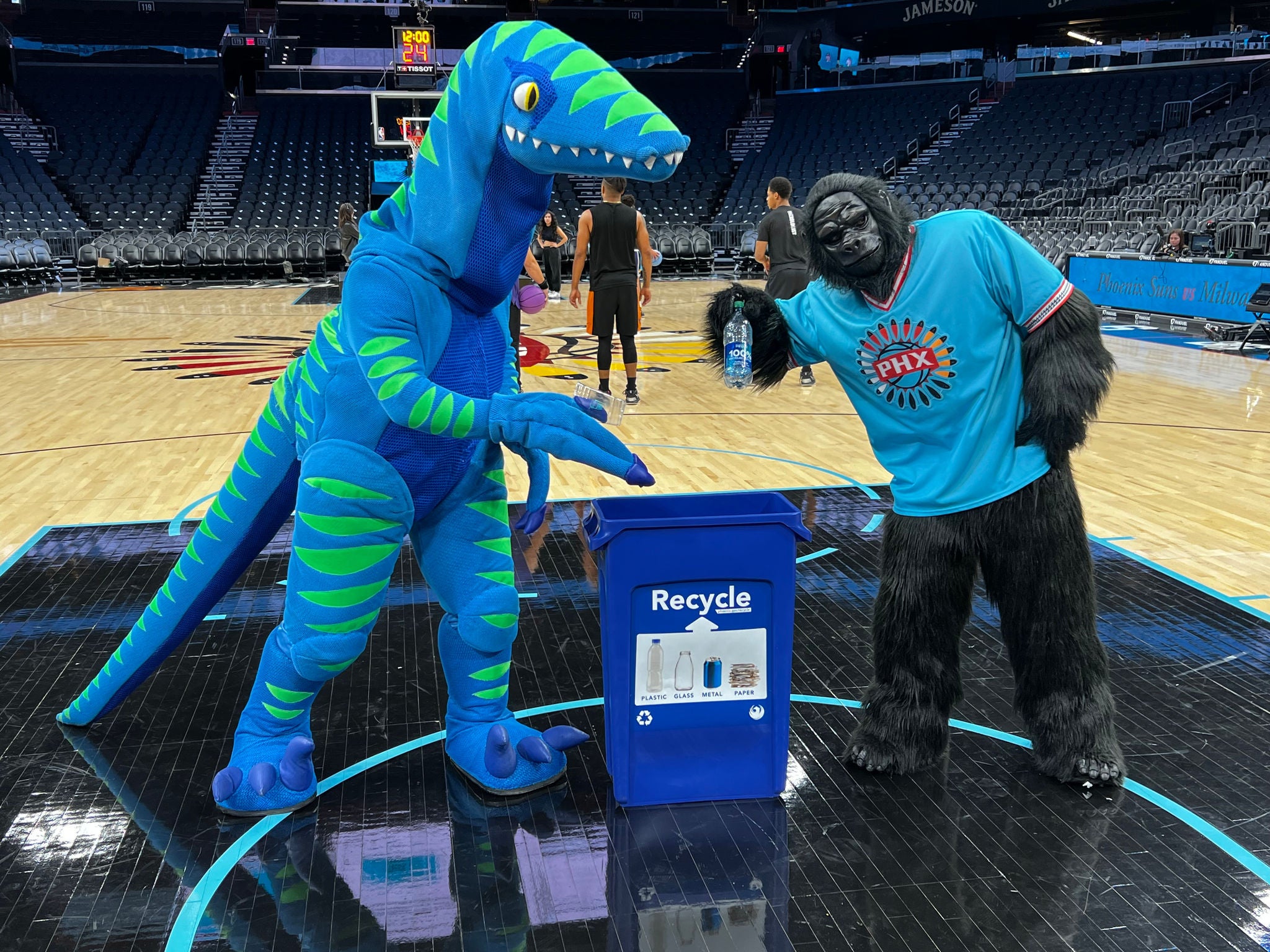 Zero, a blue and green dinosaur mascot, hangs out with the Phoenix Suns Gorilla mascot with a blue recycle bin in between them on the Suns' basketball court.