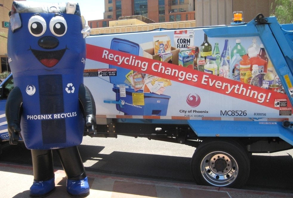 Binny, a large mascot designed to look like a blue curbside recycling container with arms and legs. He is standing next to a collection truck that says 'Recyling Changes Everything' on the side.