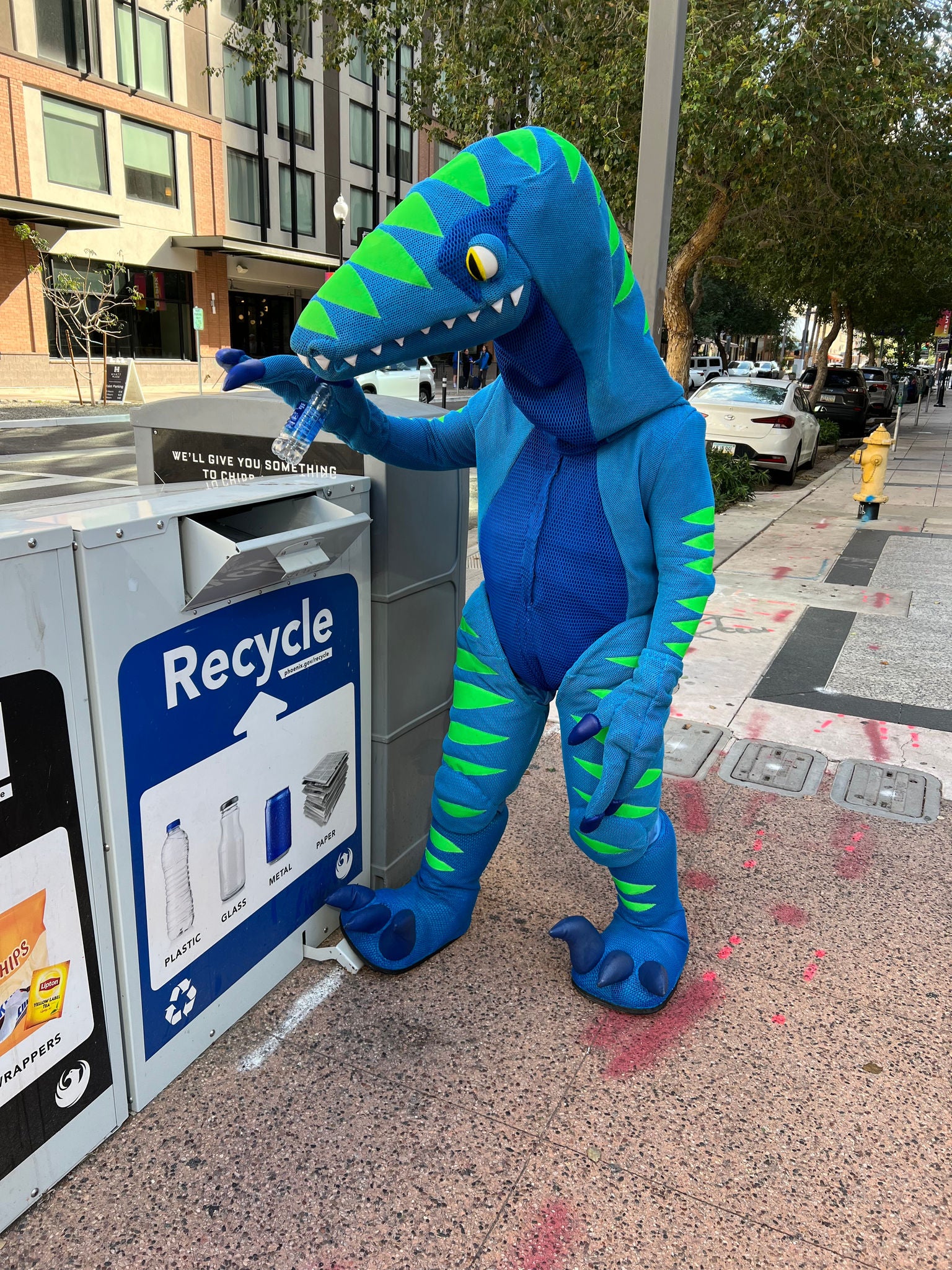 A blue and green dinosaur mascot places a plastic water bottle into a public recycle bin along a Phoenix sidewalk.