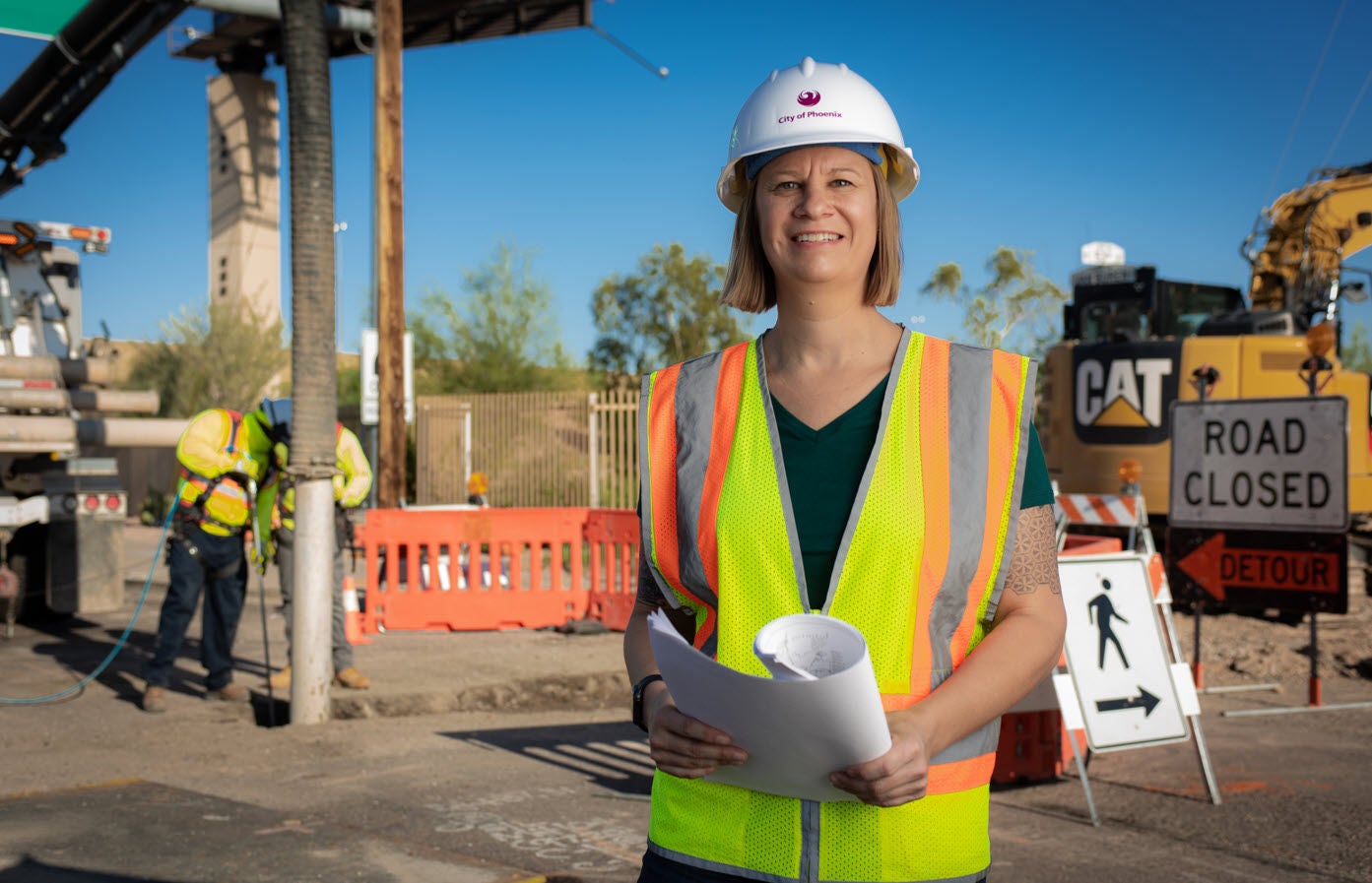 Female civil engineer standing in front of a worksite, wearing a hard hat and safety vest, and holding plans