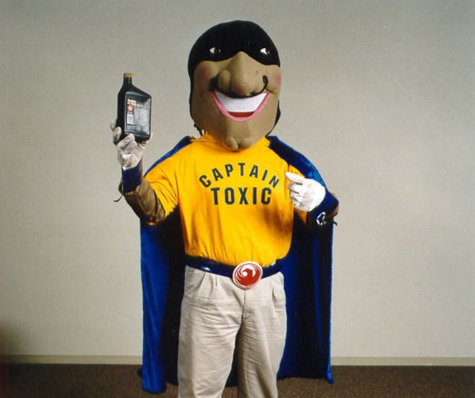 A full body shot of Captain Toxic, a costumed mascot that features an overly large head with a black mask on the eyes, a yellow shirt that says 'Captain Toxic,' a blue cape, a large belt buckle with the City of Phoenix logo, and white gloves. He is holding a bottle of motor oil, which is considered household hazardous waste.