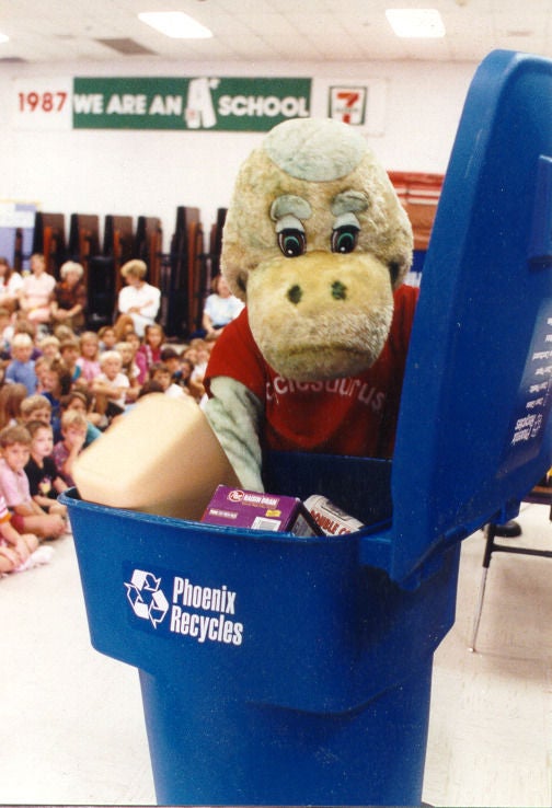 Recyclosaurus, a green dinosaur mascot with a red shirt, shows elementary school students how to properly recycling with the large blue, curbside bin.