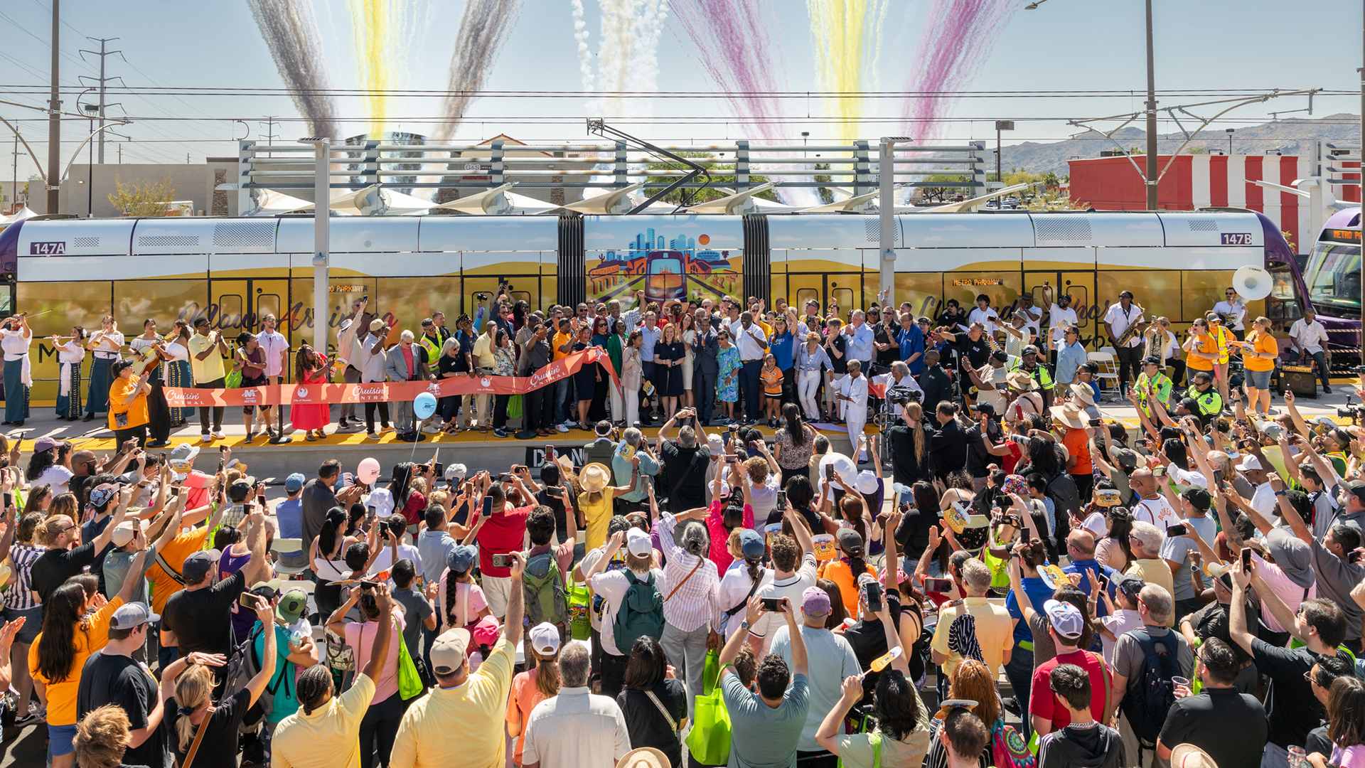 Crowd of people watching as Phoenix Mayor Kate Gallego cuts a ribbon alongside other elected officials and leaders with a light rail and fireworks are in the back. 