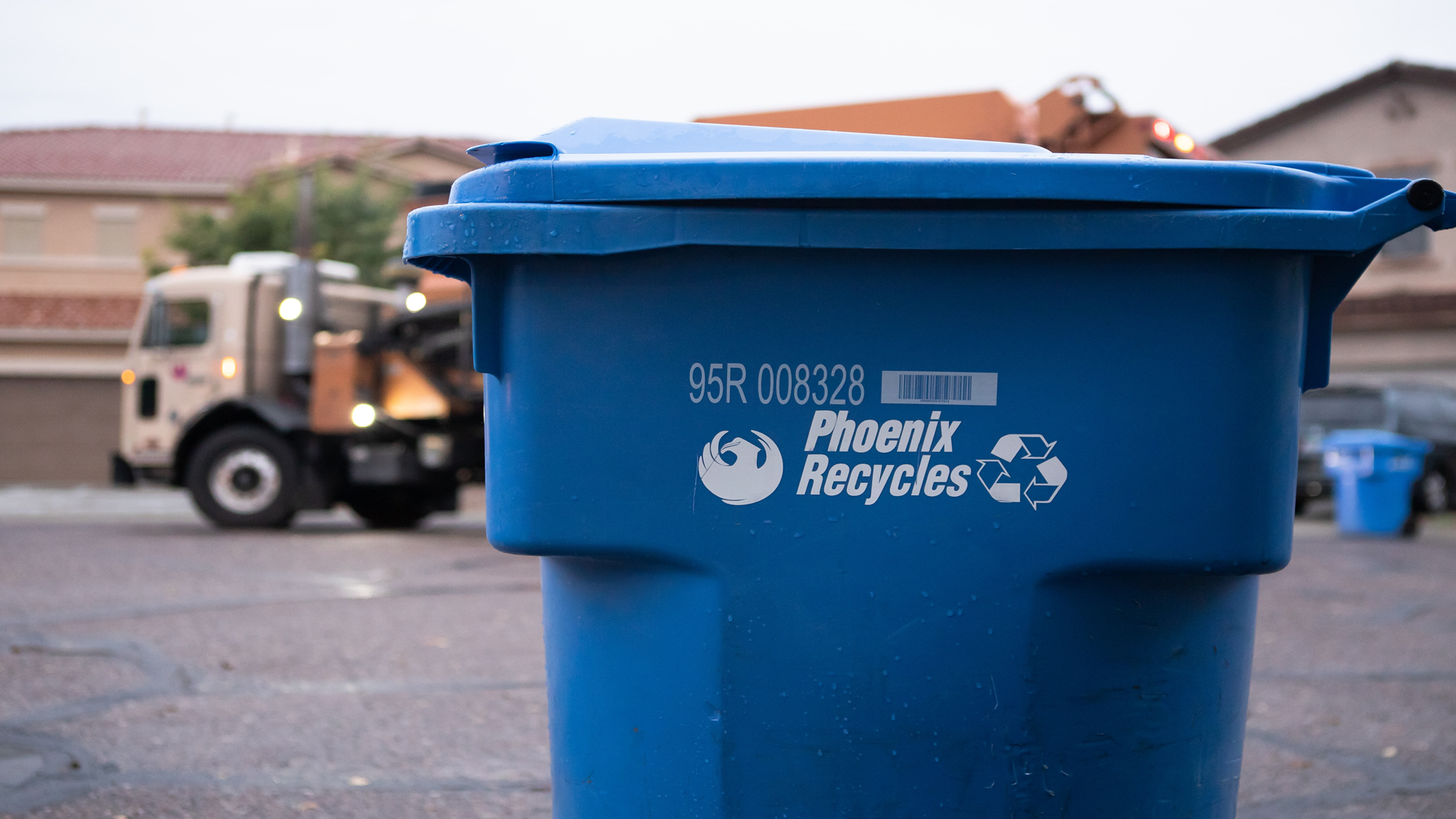 A blue curbside recycle bin that says 'Phoenix Recycles' in white letters with the City of Phoenix bird logo on one side of the text and a recycling logo (three arrows pointing at each other in a circle) on the other. In the background is a garbage truck driving down a residential street on a cloudy day.