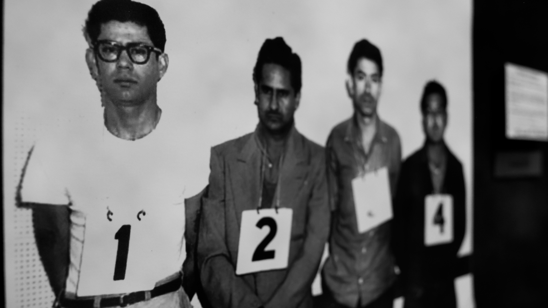 Four individuals are lined up against a plain wall. Each person is wearing a sign with a number: 1, 2, 3, and 4. The image appears to be in black and white.