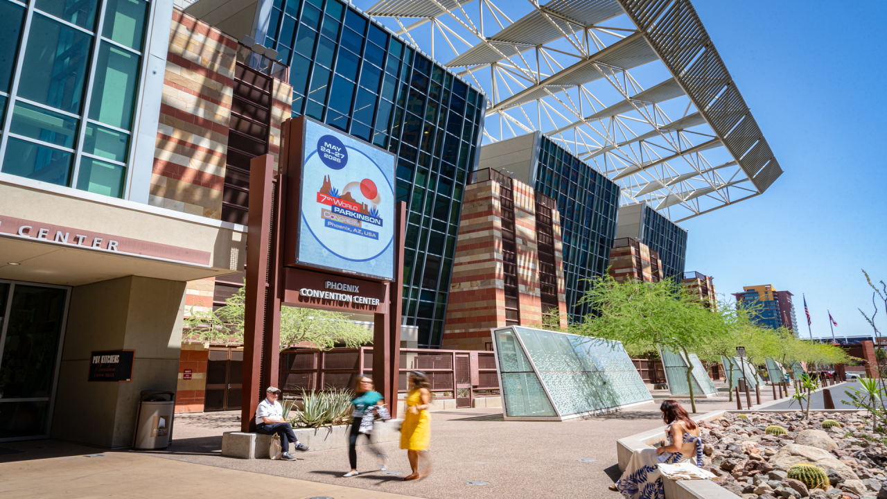 An outside shot of the Phoenix Convention Center showcases an electronic billboard that says 'May 24-27, 2026 | 7th World Parkinson Congress | Phoenix, AZ, USA. A few people are sitting and walking near the entrance to the Convention Center on a sunny day with blue, clear sky. 